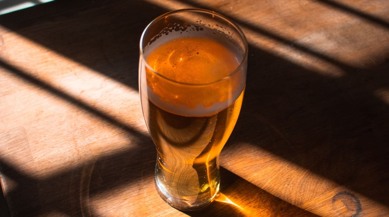 Pint glass of golden beer on a wooden table, illuminated by dramatic sunlight streaming through blinds, casting warm shadows and highlights across the surface.