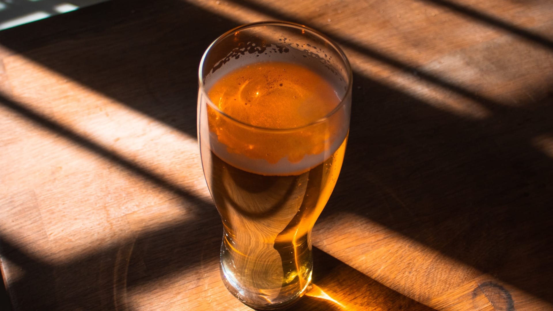Pint glass of golden beer on a wooden table, illuminated by dramatic sunlight streaming through blinds, casting warm shadows and highlights across the surface.