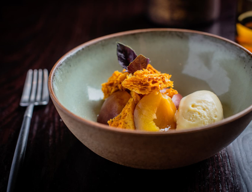 Elegant dessert in a ceramic bowl featuring honeycomb toffee, poached peaches, a scoop of vanilla ice cream, and garnished with a dark edible leaf.