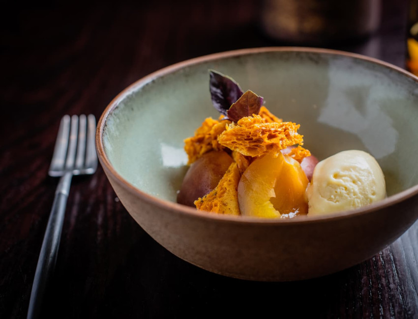 Elegant dessert in a ceramic bowl featuring honeycomb toffee, poached peaches, a scoop of vanilla ice cream, and garnished with a dark edible leaf.