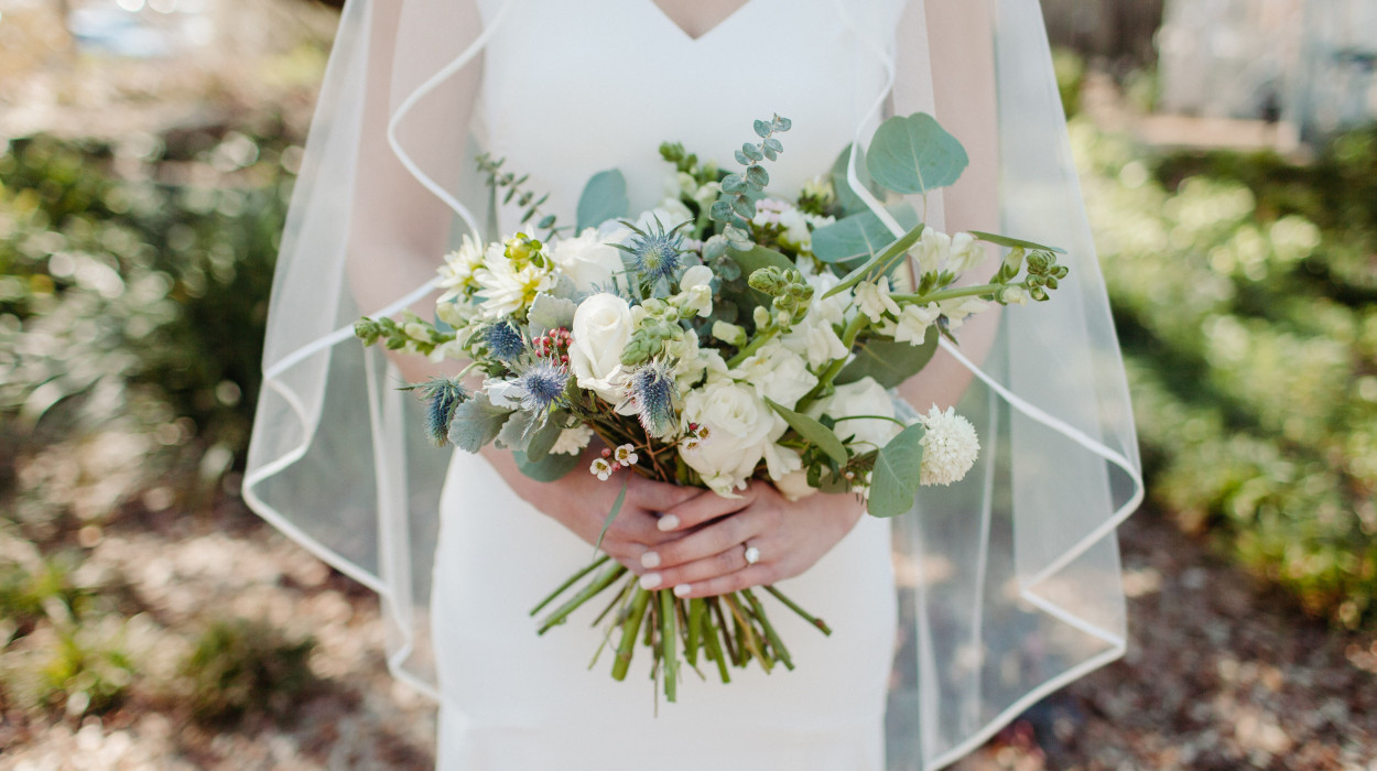 Bride in a white gown and veil holding a bouquet of white roses, greenery, and thistles, with an engagement ring visible on her finger.