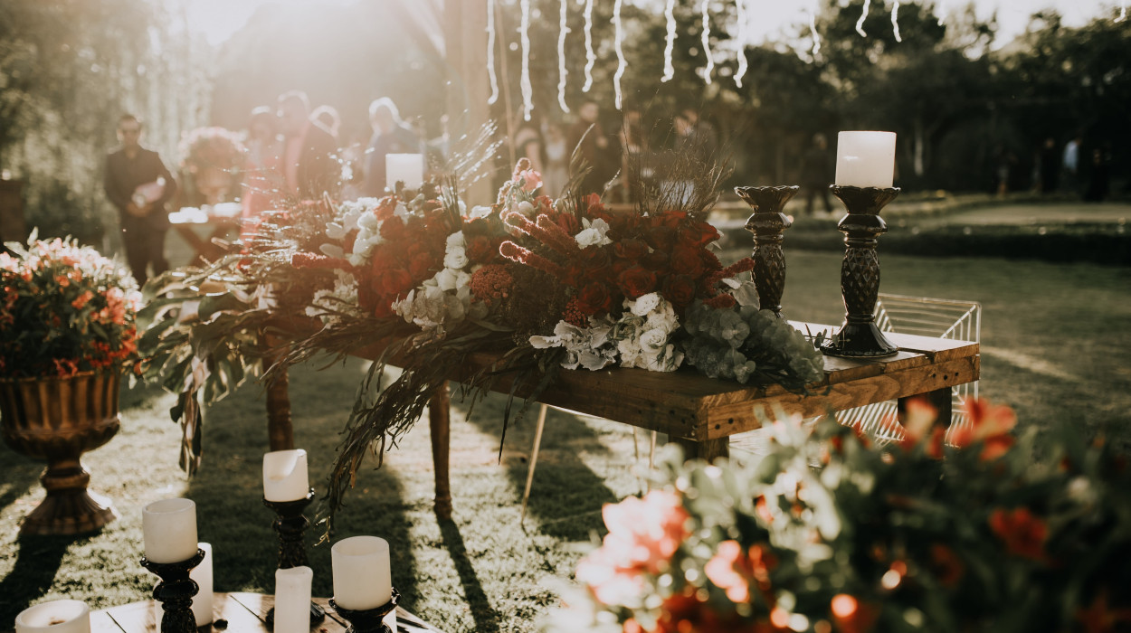 An outdoor wedding setup with a rustic wooden table adorned with red and white floral arrangements, candles, and soft sunlight filtering through the trees.