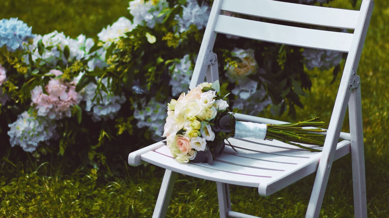 A white folding chair on grass with a bridal bouquet of white and blush roses, set beside a floral wedding arch decorated with pastel hydrangeas.