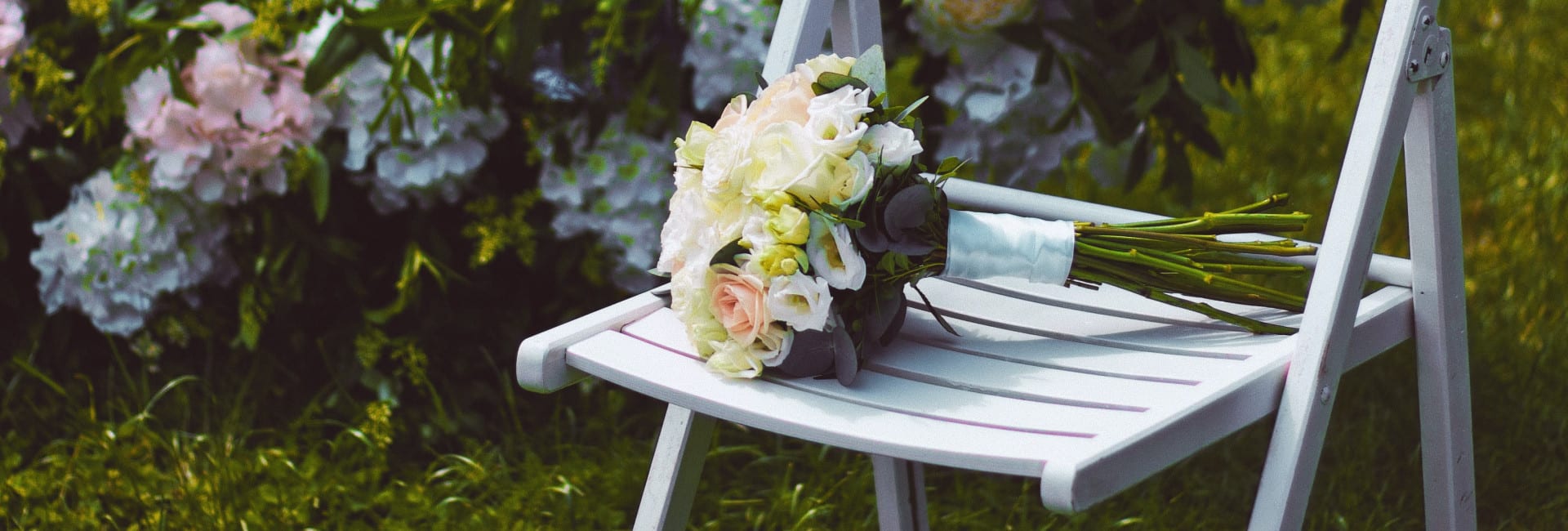 A white folding chair on grass with a bridal bouquet of white and blush roses, set beside a floral wedding arch decorated with pastel hydrangeas.