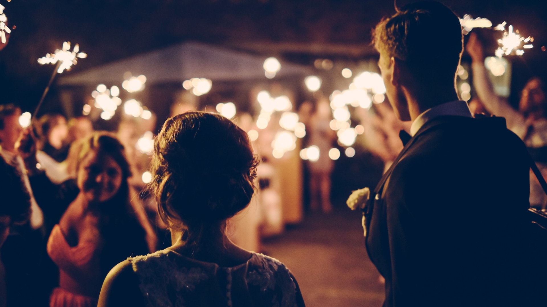 A bride and groom are seen from behind as they walk into a nighttime wedding celebration, surrounded by guests holding glowing sparklers.