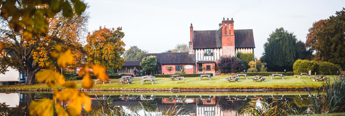 A picturesque brick and timber country house reflected in a calm pond, framed by autumn trees with picnic benches scattered across the green lawn.