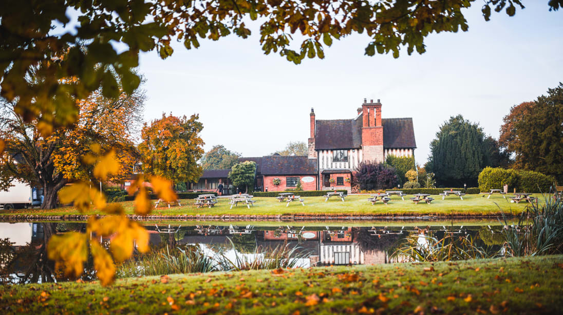 A picturesque brick and timber country house reflected in a calm pond, framed by autumn trees with picnic benches scattered across the green lawn.