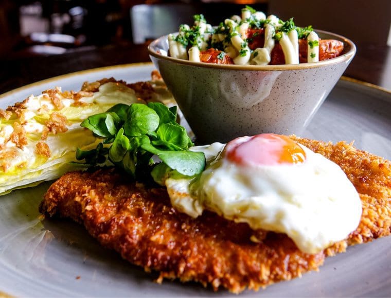 A hearty dish with crispy breaded schnitzel topped with a fried egg, fresh greens, wedge salad, and a bowl of loaded fries with sauce and herbs.