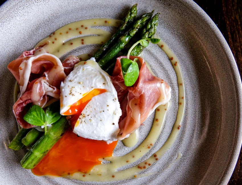 A gourmet dish featuring asparagus, prosciutto, and a poached egg with runny yolk, drizzled with mustard seed sauce on a speckled ceramic plate.
