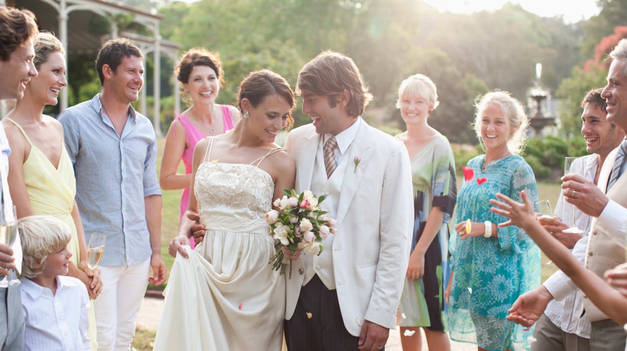 A bride and groom smile at each other while surrounded by joyful wedding guests outdoors, with the bride holding a bouquet and wearing a white dress.