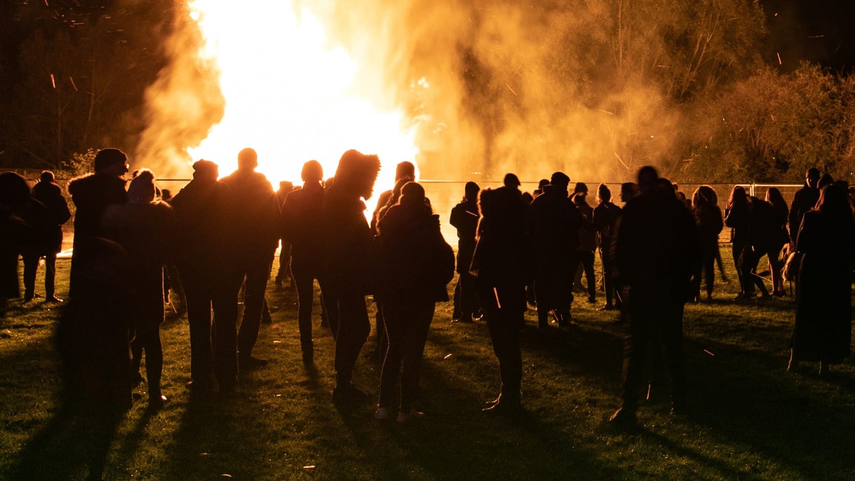 A large circle of people stands around a crackling bonfire, creating a warm and inviting scene of camaraderie and fun.