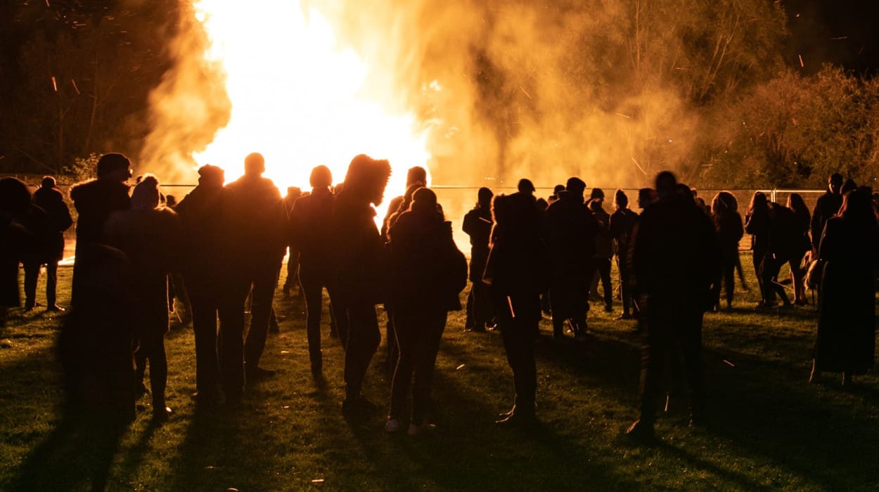 A large circle of people stands around a crackling bonfire, creating a warm and inviting scene of camaraderie and fun.