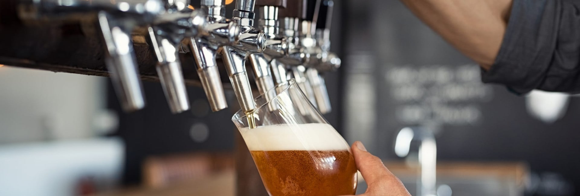 A person fills a glass with beer from a tap at a bar, holding the glass at an angle to create a foamy head. Several beer taps are visible in the background.