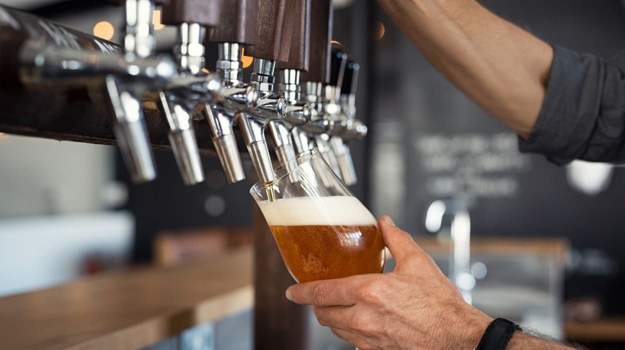 A person fills a glass with beer from a tap at a bar, holding the glass at an angle to create a foamy head. Several beer taps are visible in the background.
