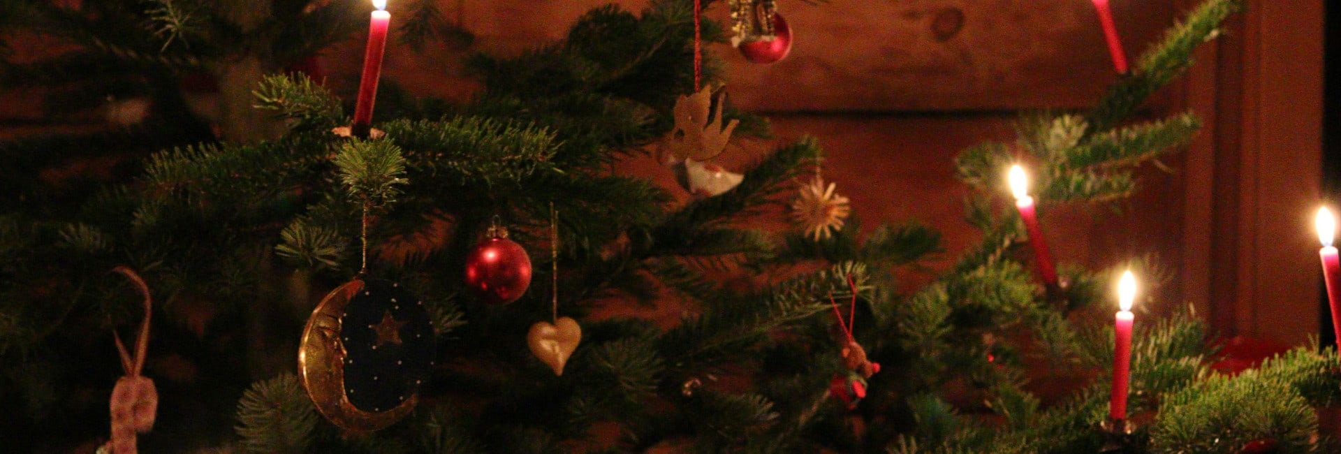 A Christmas tree decorated with red baubles, ornaments, and lit red candles, creating a warm glow in a cozy wooden room.