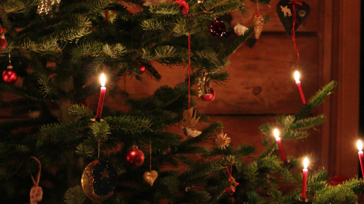 A Christmas tree decorated with red baubles, ornaments, and lit red candles, creating a warm glow in a cozy wooden room.