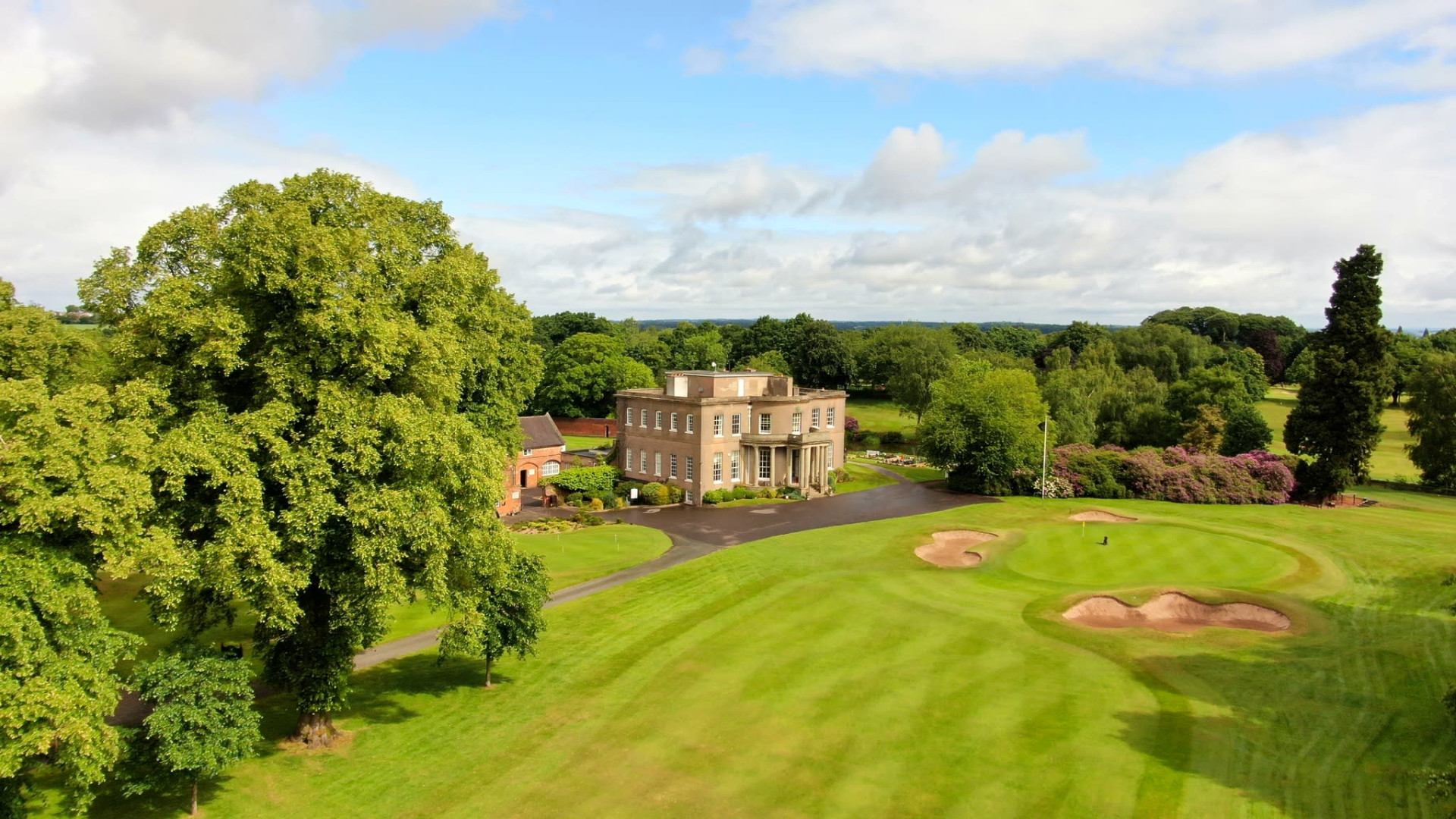 Aerial view of a stately country house beside a well-manicured golf course with sand bunkers, surrounded by lush green trees under a partly cloudy sky.