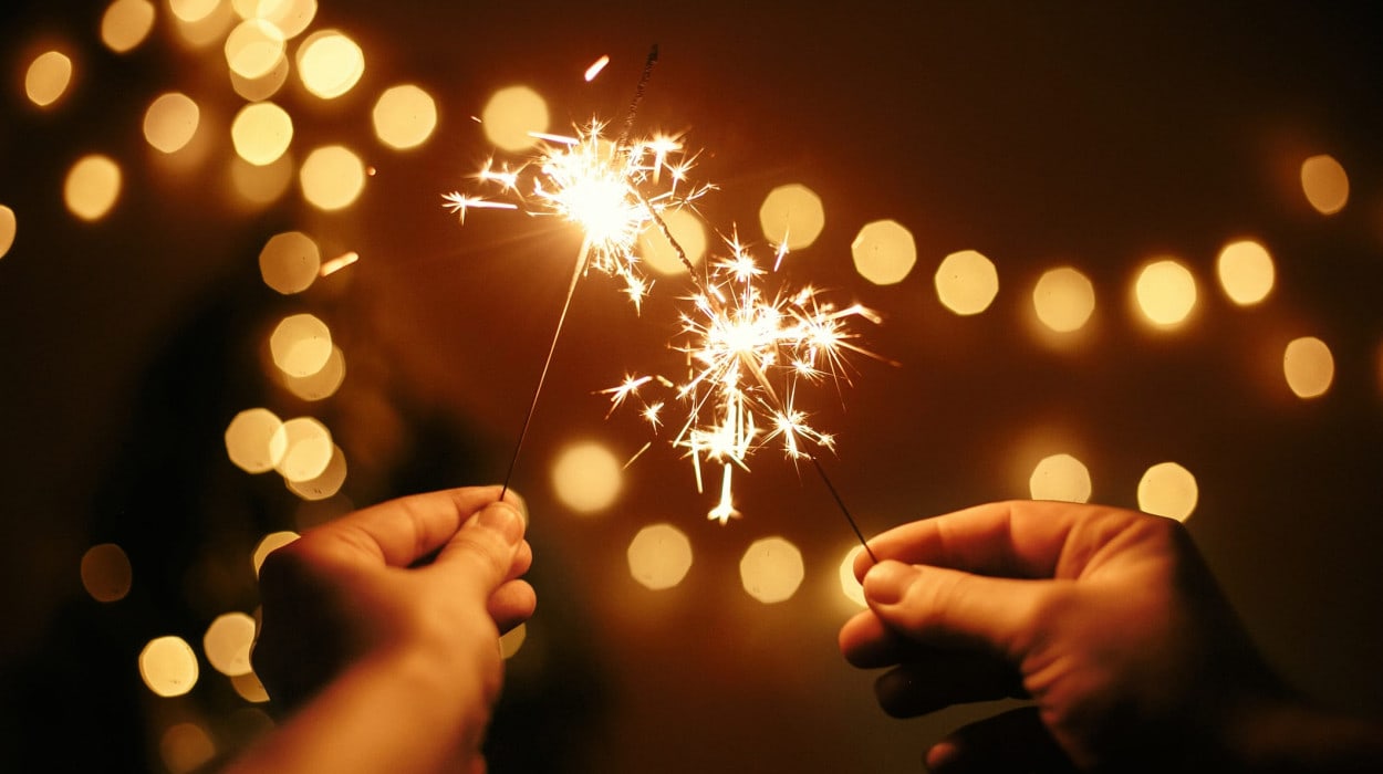 Two people holding sparklers with glowing sparks, set against a warm, blurry background of golden lights, creating a festive and celebratory atmosphere.