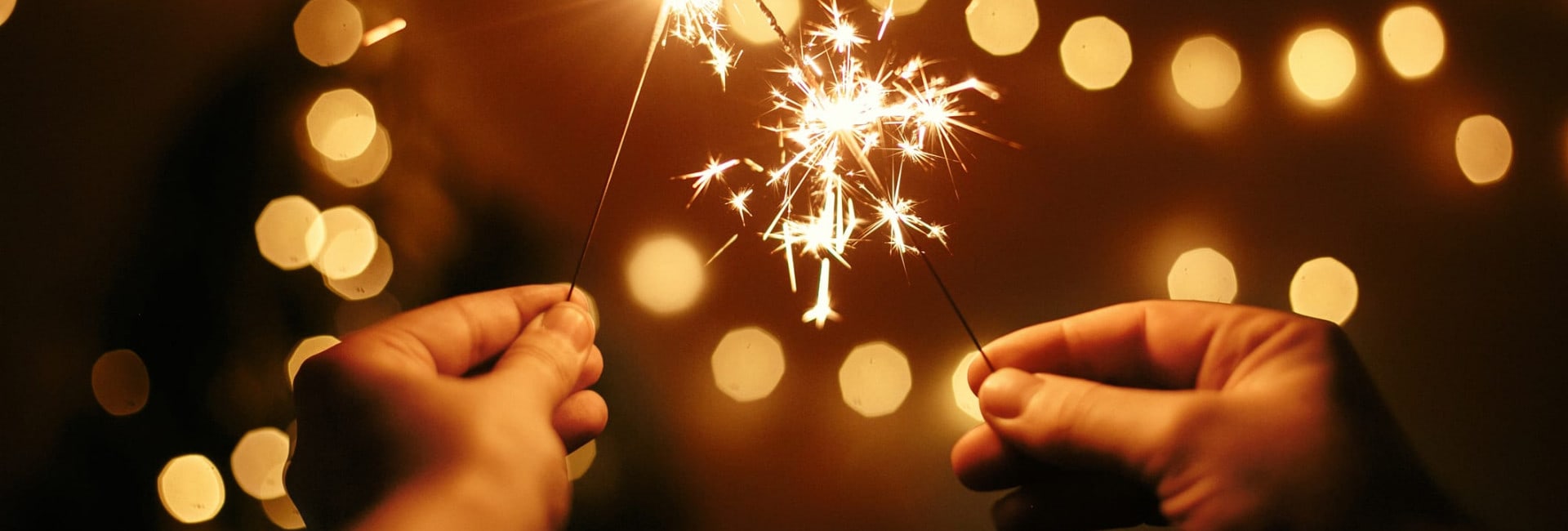 Two people holding sparklers with glowing sparks, set against a warm, blurry background of golden lights, creating a festive and celebratory atmosphere.