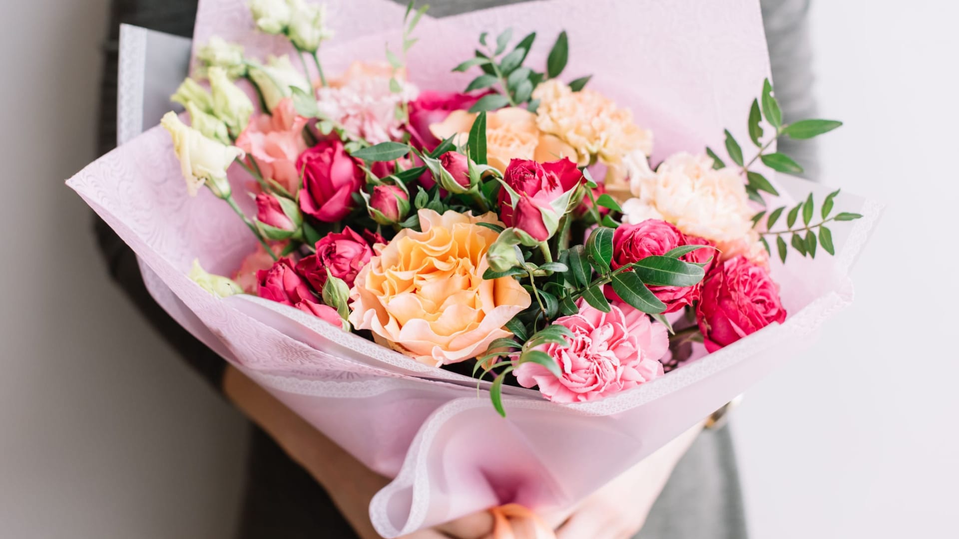 A person in a gray outfit holds a bouquet of pink, red, and peach roses and carnations, wrapped in soft pink paper with greenery accents.