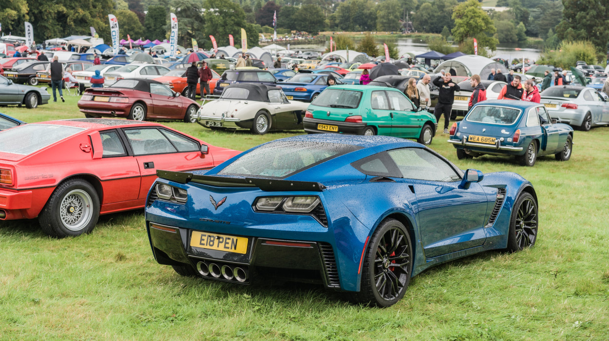 A blue Chevrolet Corvette with a wet exterior is parked on a grassy field at a classic car show, surrounded by vintage cars and a crowd under an overcast sky.