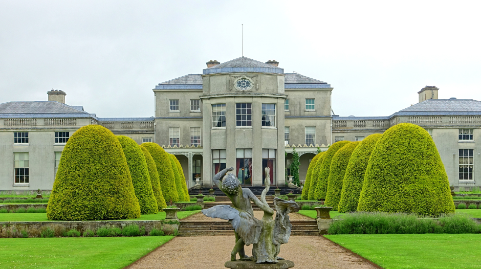 A grand estate with manicured topiary trees, a central statue fountain, and a large neoclassical mansion in the background under a cloudy sky.