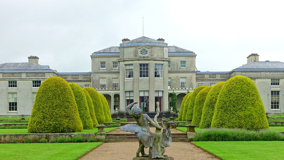 A grand estate with manicured topiary trees, a central statue fountain, and a large neoclassical mansion in the background under a cloudy sky.