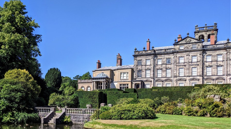 A grand historic mansion with formal gardens and a pond filled with lily pads, set against a clear blue sky and surrounded by lush greenery.