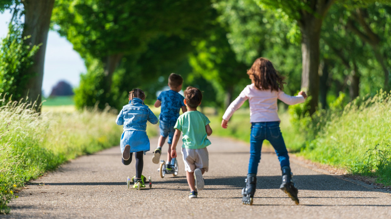 Four children riding scooters and rollerblading down a tree-lined path on a sunny day, surrounded by greenery and tall grass.