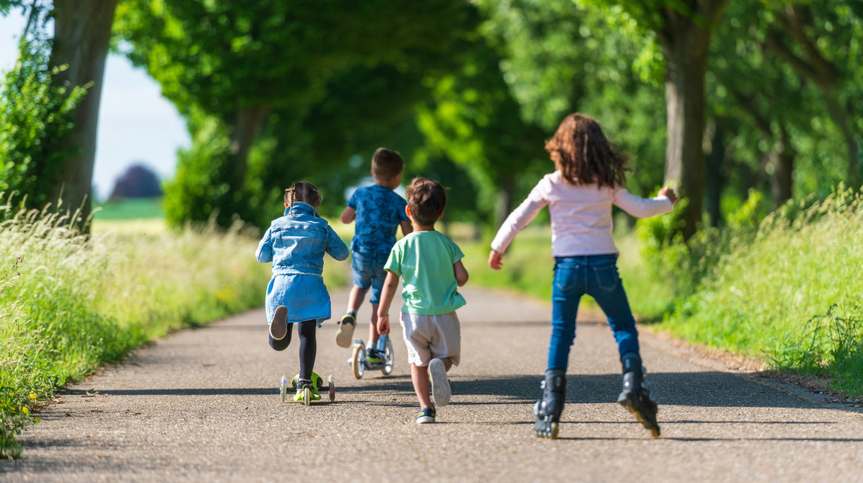 Four children riding scooters and rollerblading down a tree-lined path on a sunny day, surrounded by greenery and tall grass.