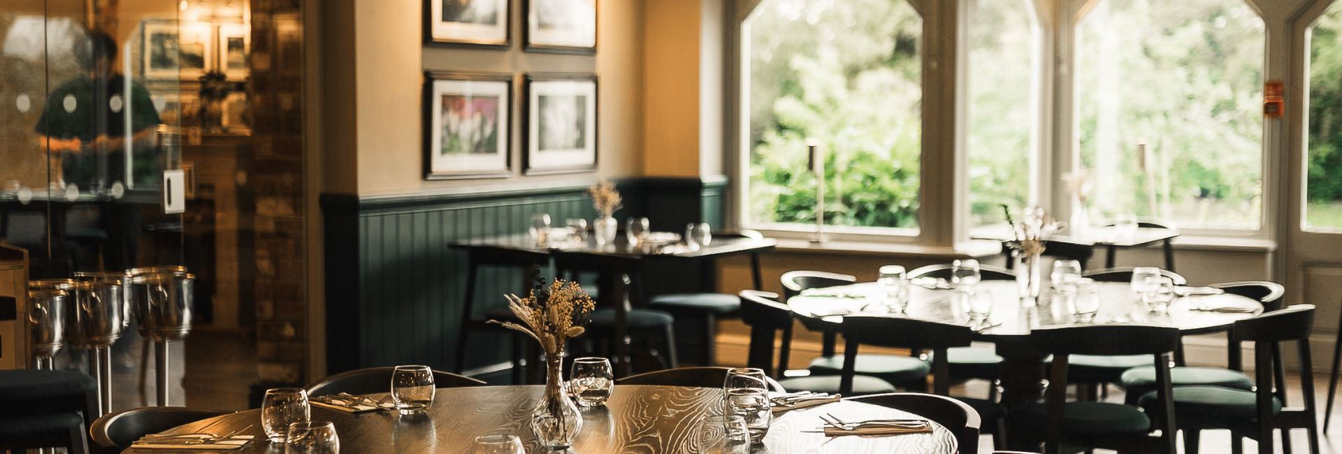 Bright restaurant space with light streaming in from tall windows, wooden floors, round tables set with glassware and dried floral centerpieces.